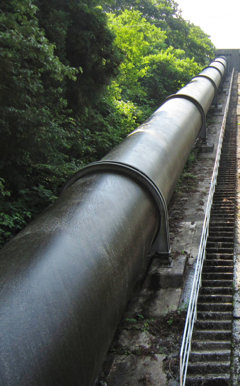 Metal penstock moving downhill outdoors with green trees in the background.